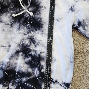 May include: A tie-dye hoodie in black and white with the word "ME" printed on the front. The hoodie has a drawstring hood and a front pocket. A ruler is placed next to the hoodie.