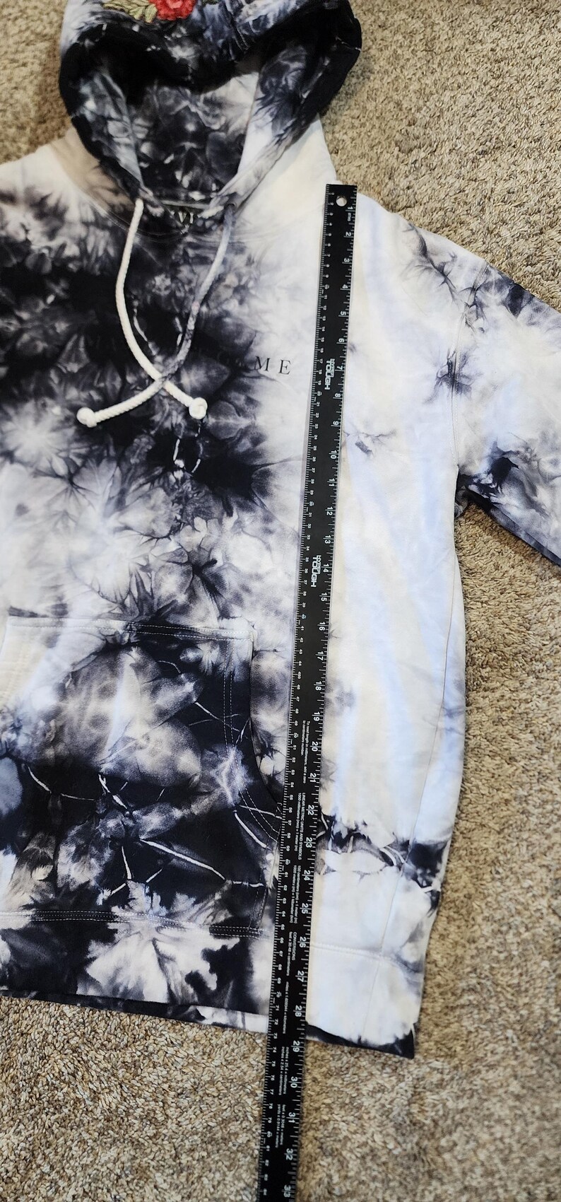May include: A tie-dye hoodie in black and white with the word "ME" printed on the front. The hoodie has a drawstring hood and a front pocket. A ruler is placed next to the hoodie.
