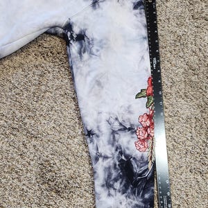 May include: A tie-dye hoodie in shades of white, black, and gray. The sleeve features a floral design with red and pink flowers and green leaves. The word "ME" is visible on the upper part of the hoodie.