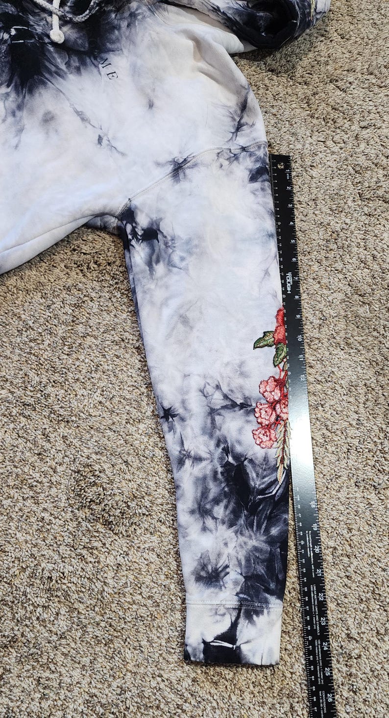 May include: A tie-dye hoodie in shades of white, black, and gray. The sleeve features a floral design with red and pink flowers and green leaves. The word "ME" is visible on the upper part of the hoodie.