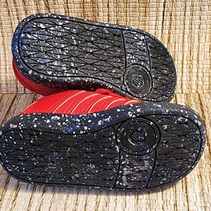 May include: Red and white athletic shoes with a black sole featuring white and blue speckles. The soles have a textured pattern and the brand logo is visible. The shoes are designed for sports and casual wear.