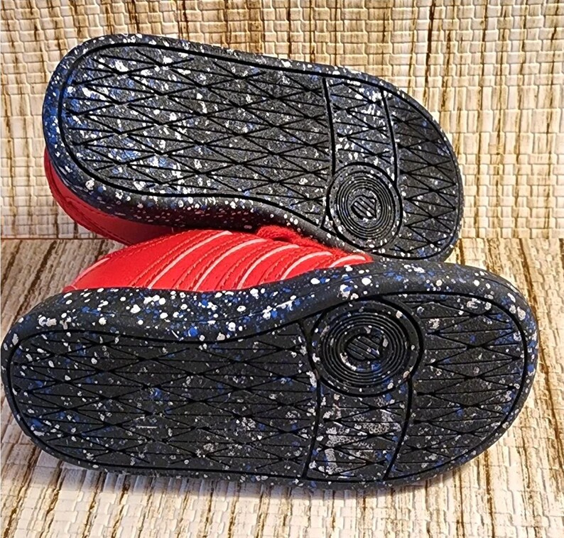 May include: Red and white athletic shoes with a black sole featuring white and blue speckles. The soles have a textured pattern and the brand logo is visible. The shoes are designed for sports and casual wear.