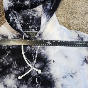 May include: A tie-dye hoodie in black and white with a drawstring hood. The word "ME" is printed on the front. A ruler is placed across the hoodie. A red rose is embroidered on the sleeve.
