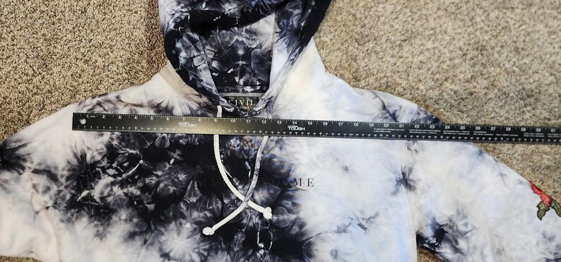 May include: A tie-dye hoodie in black and white with a drawstring hood. The word "ME" is printed on the front. A ruler is placed across the hoodie. A red rose is embroidered on the sleeve.