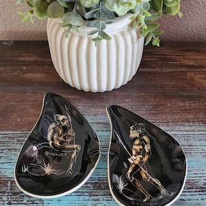 May include: Two black, teardrop-shaped decorative dishes with white rims. Each dish features a gold illustration of a figure and the word "SYDNEY". A white, ribbed ceramic pot with greenery sits in the background.