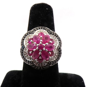 May include: A silver ring featuring a cluster of oval-cut vibrant pink gemstones at the center, surrounded by a border of small, dark, shimmering stones set in a scalloped, flower-like frame with a polished metallic finish and intricate detailing.