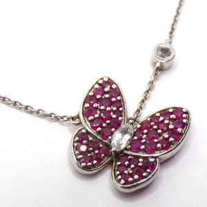 May include: A silver-toned necklace featuring a butterfly-shaped pendant. The butterfly wings are encrusted with numerous small, round, deep pink stones, while the center body is a single, clear, marquise-cut stone. A small, round, clear stone is set on the chain.
