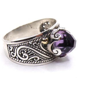 May include: A silver ring with an ornate design and a large, faceted amethyst gemstone. The ring has a wide band with intricate details.