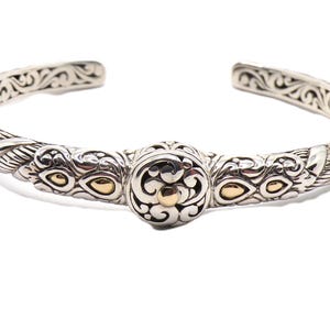 May include: Silver cuff bracelet with ornate detailing and gold accents. The bracelet features a twisted rope design on the sides and a central decorative element. The open design allows for easy wear. A piece of jewelry.