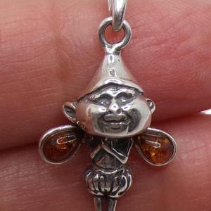 May include: A silver pendant shaped like a small figure wearing a pointed hat and puffy shorts, featuring two teardrop-shaped amber stones on either side of the torso. The figure has a detailed face and is attached to a silver bail at the top.