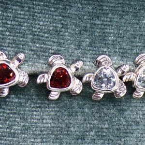 May include: Four silver turtle-shaped stud earrings. Two have red gemstones, and two have blue gemstones. The earrings are displayed on a teal green velvet surface.