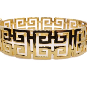 Puede incluir: Una pulsera dorada con un patrón geométrico repetitivo. La pulsera tiene un cierre de bisagra y una serie de cuadrados y rectángulos calados. El diseño es de estilo clásico y atemporal.