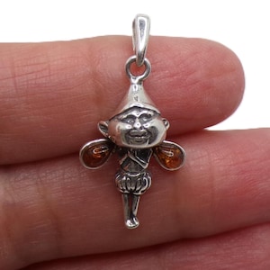 Puede incluir: Un colgante de plata con forma de hada o elfo caprichoso. El colgante presenta una cara sonriente, un gorro puntiagudo y alas de color ámbar. El hada está representada con detalles y un diseño juguetón.