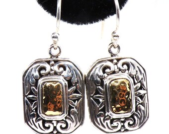 May include: A pair of silver-tone earrings with a gold-tone rectangular center. The earrings have an ornate, filigree design around the gold center. The earrings are hanging from a black surface.