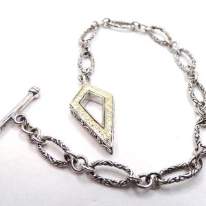 May include: A silver-toned bracelet with an ornate chain design and a diamond-shaped pendant. The pendant features a cream-colored center and is accented with small, sparkling stones. The bracelet has a T-bar clasp.