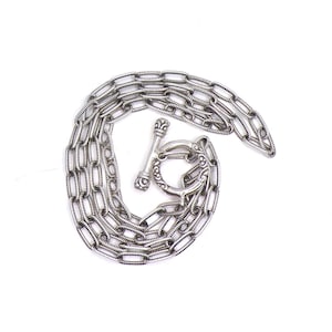 May include: Silver-toned necklace with a toggle clasp. The chain is composed of rectangular links, and the clasp features decorative detailing. The necklace is displayed against a white background.