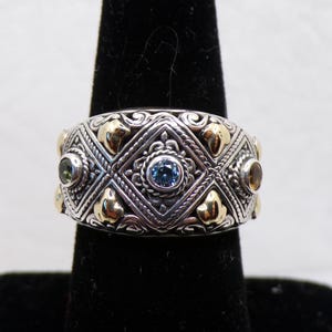 May include: Silver ring with gold accents and gemstones. The ring features a central blue gemstone, surrounded by intricate silver filigree and gold heart-shaped details. Additional gemstones are set on the sides.