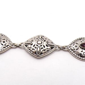 May include: Silver bracelet featuring ornate, diamond-shaped links with filigree detailing. Each link is accented with a small, round, dark red gemstone. The bracelet has a toggle clasp for secure fastening. A piece of jewelry.