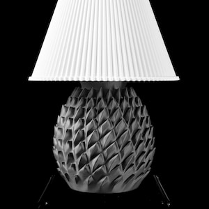 Può includere: Lampada in bianco e nero con base a forma di ananas e paralume bianco plissettato. La base presenta un design geometrico strutturato. Lo sfondo è nero.