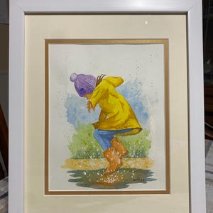 May include: Watercolor painting in a white frame. The artwork depicts a child in a yellow raincoat and purple hat jumping in a puddle. The child wears blue jeans and brown rain boots. The background features green grass and blue sky.
