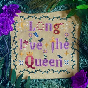 Puede incluir: Bordado de punto de cruz con fondo amarillo y enredaderas verdes. El texto dice "Long Live the Queen" en letras moradas y rojas. El diseño incluye una corona, una espada, hongos, pájaros y flores.