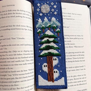 May include: A blue cross-stitch bookmark depicting a winter scene with a full moon, snow-covered trees, and a cabin. The bookmark is in an open book.