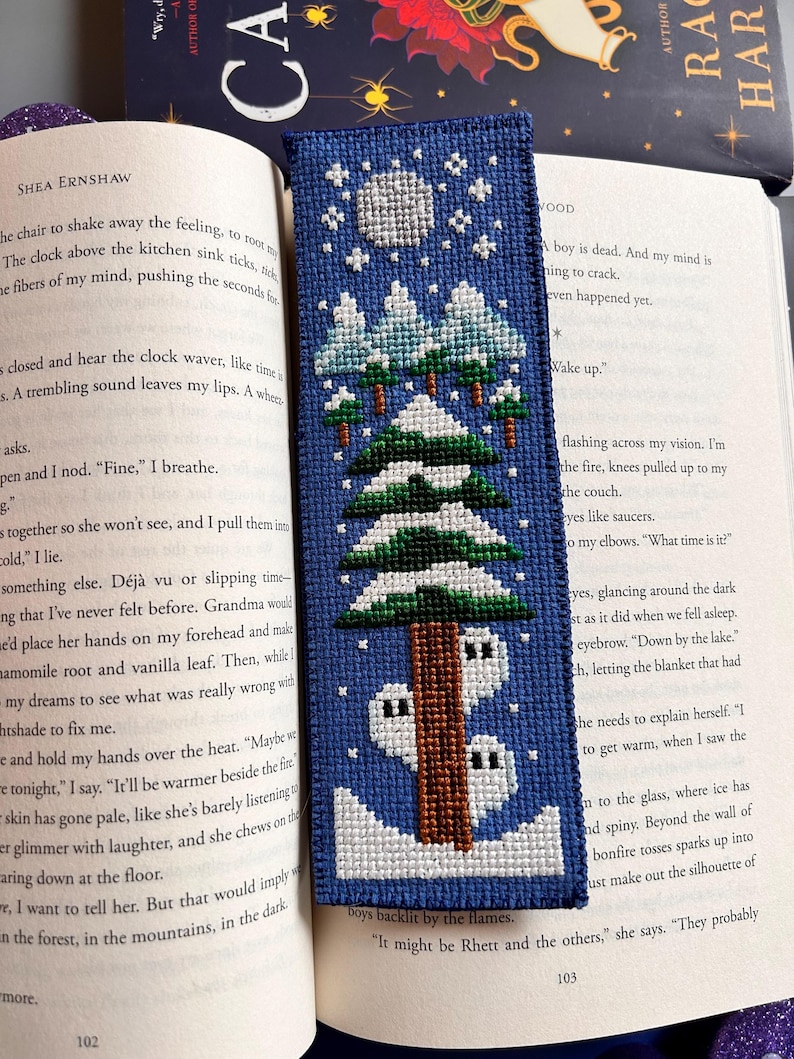 May include: A blue cross-stitch bookmark depicting a winter scene with a full moon, snow-covered trees, and a cabin. The bookmark is in an open book.