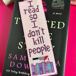 May include: A handmade pink cross-stitch bookmark with the text "I read so I don't kill people." The bookmark features a knife and blood design at the bottom. The bookmark is placed on top of a book.
