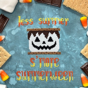 May include: A cross-stitch embroidery on blue fabric featuring a marshmallow with a jack-o'-lantern face between two graham crackers. The text reads less summer, S'MORE, and SUMMERWEEN in orange and yellow gradient thread, surrounded by candy corn.