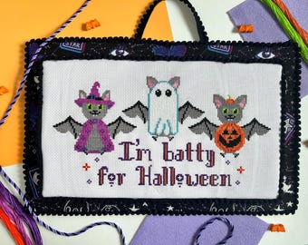 May include: Cross-stitch Halloween wall hanging featuring three bats: a witch bat, a ghost bat, and a pumpkin bat. The phrase "I'm batty for Halloween" is stitched below. The border is black with a scalloped edge and a black handle.