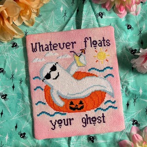 May include: A pink cross-stitch design with the words "Whatever floats your ghost." It depicts a ghost in sunglasses on an orange inflatable, with a yellow sun and blue waves. The design is on a teal background.