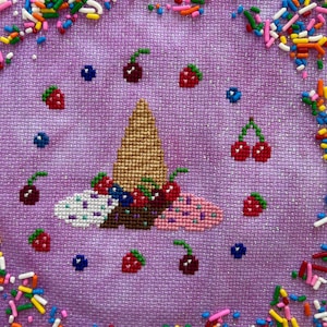 May include: Cross-stitch design of an ice cream cone with a waffle cone, two scoops, and fruit toppings. The background is a lavender color with scattered berries and sprinkles. The border is made of colorful sprinkles.