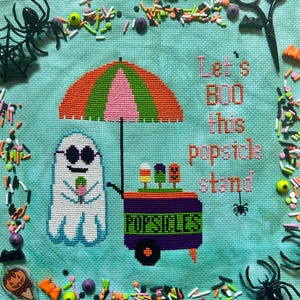 May include: A cross-stitch pattern on light blue fabric featuring a white ghost wearing sunglasses, holding a popsicle next to a purple popsicle stand under a striped umbrella. The text reads Let's BOO this popsicle stand. Surrounded by plastic spiders.