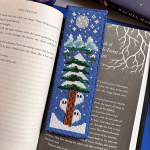May include: A blue cross-stitch bookmark depicting a winter scene with snow-covered trees, a full moon, and a small house. The bookmark is in an open book. The word "CACKLE" is visible on a book spine. The book is open to page 39.