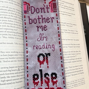 May include: A handmade bookmark with the text "Don't bother me I'm reading OR ELSE" in black and red cross-stitch. The bookmark is gray with a red border and is placed on top of an open book.