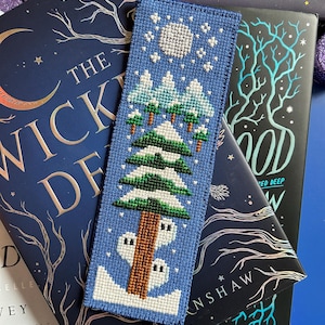 May include: A blue cross-stitch bookmark depicting a winter scene with a full moon, snow-covered trees, and a house. The bookmark rests on a book with a dark blue cover and gold lettering. Purple candle holders are in the background.