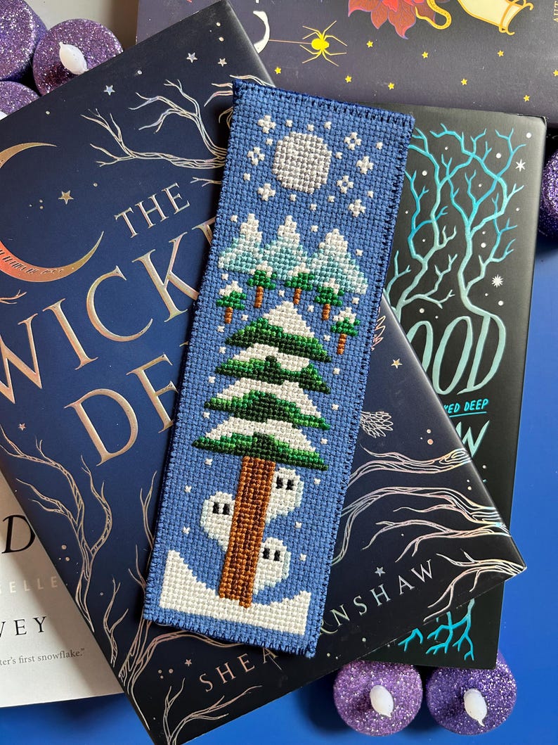 May include: A blue cross-stitch bookmark depicting a winter scene with a full moon, snow-covered trees, and a house. The bookmark rests on a book with a dark blue cover and gold lettering. Purple candle holders are in the background.