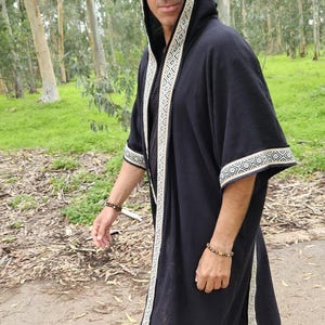 May include: Black hooded robe with white geometric trim. The robe has long sleeves and a flowing design. The trim is a repeating pattern. The person is wearing sunglasses and a bracelet. The background is a natural outdoor setting.