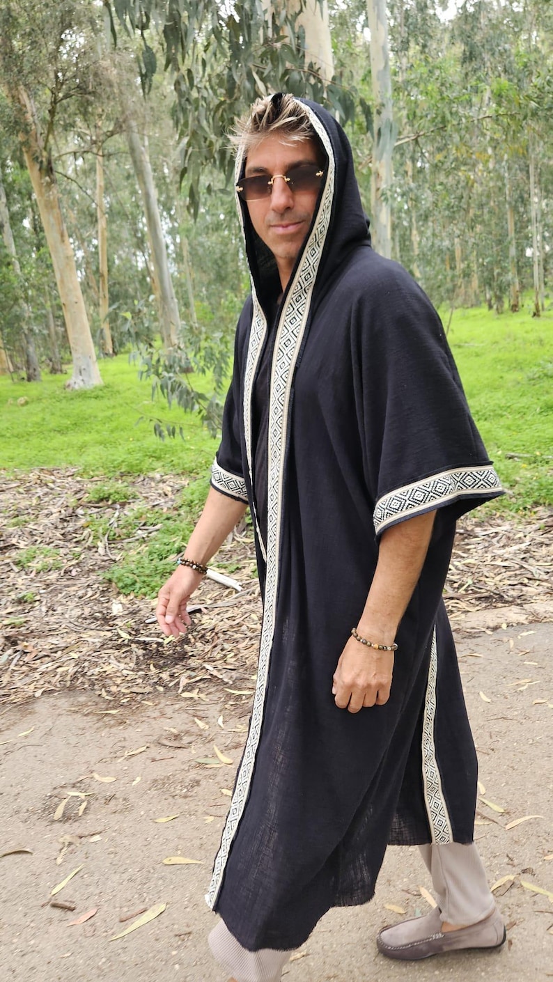 May include: Black hooded robe with white geometric trim. The robe has long sleeves and a flowing design. The trim is a repeating pattern. The person is wearing sunglasses and a bracelet. The background is a natural outdoor setting.