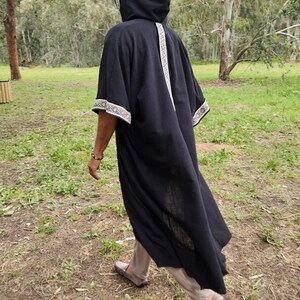 May include: A black hooded robe with white embroidered trim on the hood, sleeves, and down the back. The robe is worn over beige trousers and brown shoes. The person is walking on a grassy path.