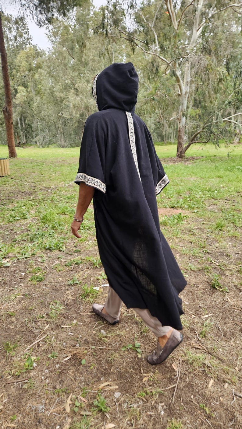 May include: A black hooded robe with white embroidered trim on the hood, sleeves, and down the back. The robe is worn over beige trousers and brown shoes. The person is walking on a grassy path.