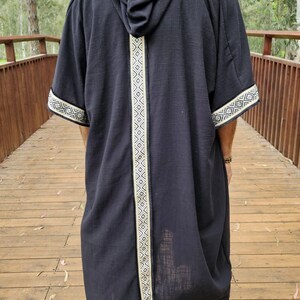May include: A black hooded robe with white geometric trim along the hood, centre, and sleeves. The robe is worn over light-coloured trousers and shoes. The person is standing on a wooden bridge.