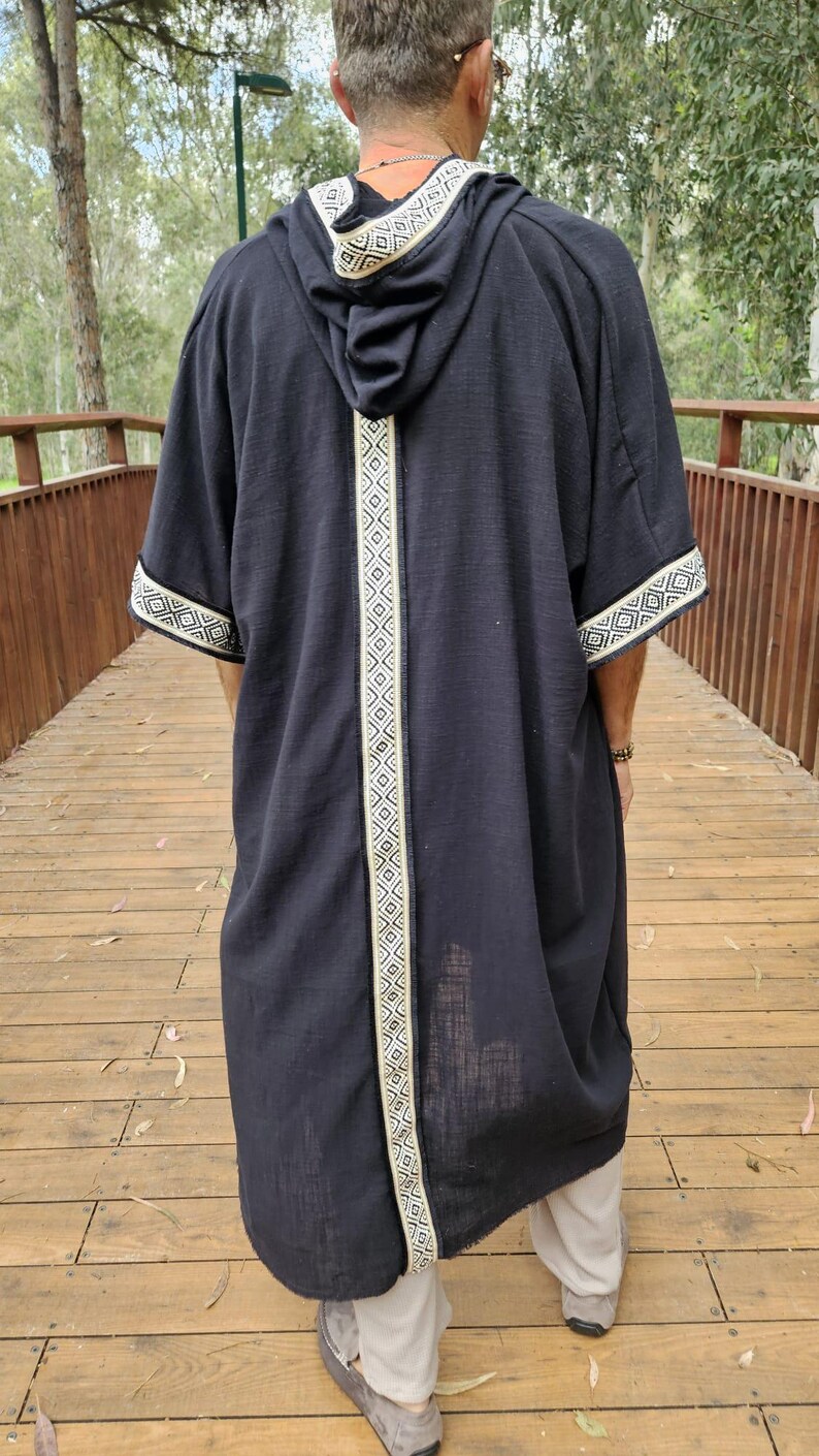 May include: A black hooded robe with white geometric trim along the hood, centre, and sleeves. The robe is worn over light-coloured trousers and shoes. The person is standing on a wooden bridge.