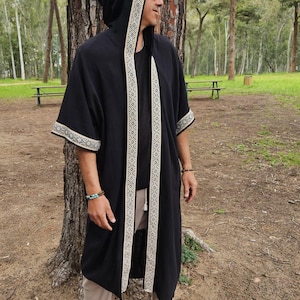 May include: Black hooded robe with cream geometric trim on the hood, front, and sleeves. The robe is worn over a black top and beige trousers. The person is wearing brown loafers and bracelets. The background is a park with trees and benches.