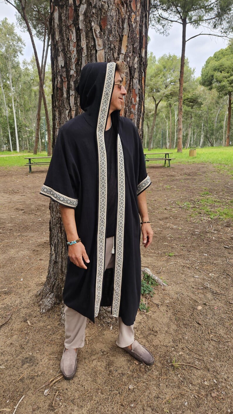 May include: Black hooded robe with cream geometric trim on the hood, front, and sleeves. The robe is worn over a black top and beige trousers. The person is wearing brown loafers and bracelets. The background is a park with trees and benches.