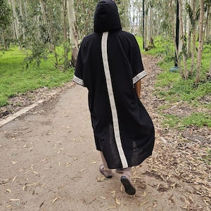 May include: A black hooded robe with white patterned trim and a white stripe down the back. The person is walking on a path in a forest. The robe is knee-length.