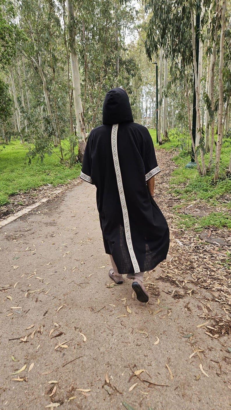 May include: A black hooded robe with white patterned trim and a white stripe down the back. The person is walking on a path in a forest. The robe is knee-length.