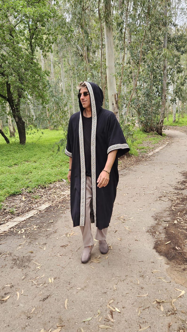 May include: A black hooded robe with white and beige trim, worn over a black shirt and beige trousers. The robe is long and flowing, with a hood and decorative trim along the edges. The person is walking on a dirt path in a wooded area.