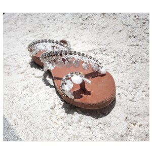 White leather sandals with a white lace and rhinestone embellishment. The sandals have a brown leather sole.