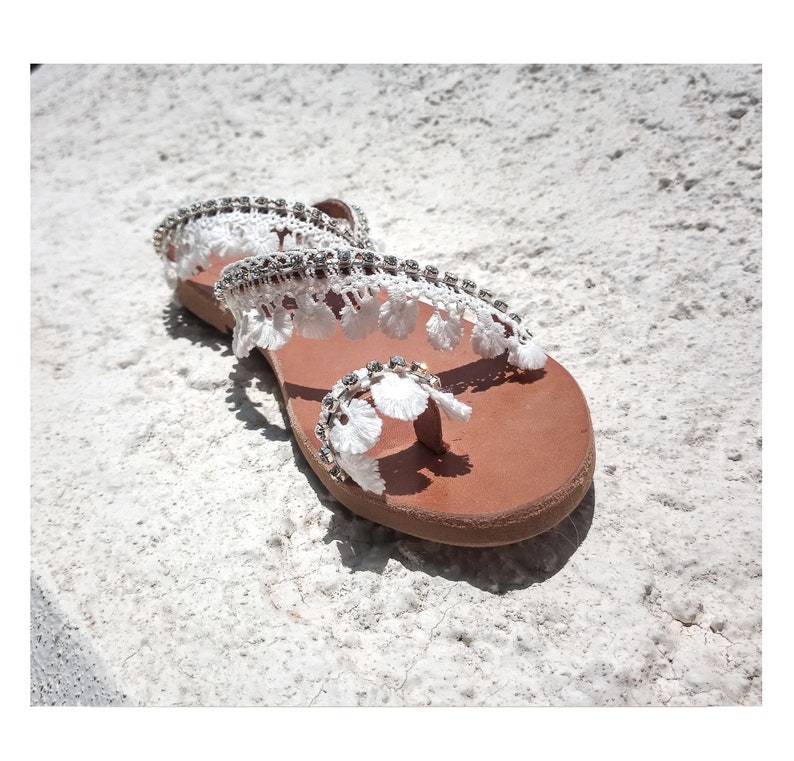 White leather sandals with a white lace and rhinestone embellishment. The sandals have a brown leather sole.
